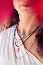 Load image into Gallery viewer, Emily Koliandri in a white dress is wearing a combo in blue tones of 5 necklaces made of japanese miyuki beads, silver 925 gold plated chains, combined with silver 925 gold plated round, rectangular and triangle charms with messages.

